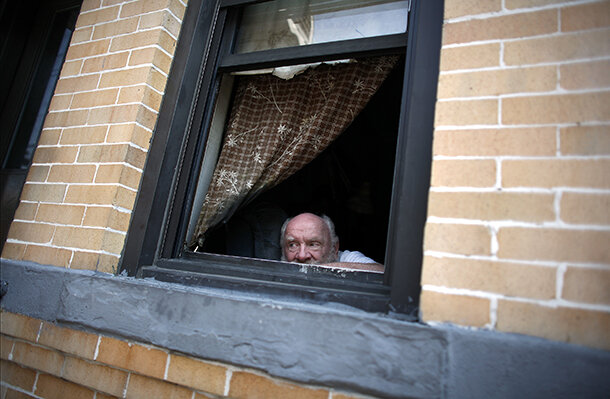 A man looks out a window from an apartment in a brick building 
