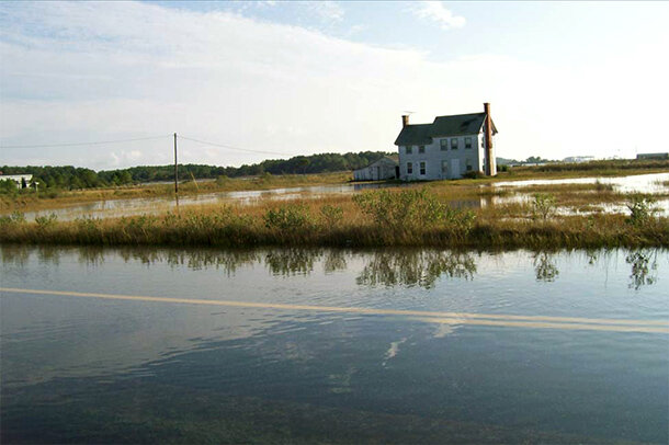Abandoned house and shed surrounded by open water and marsh near a flooded two-lane road