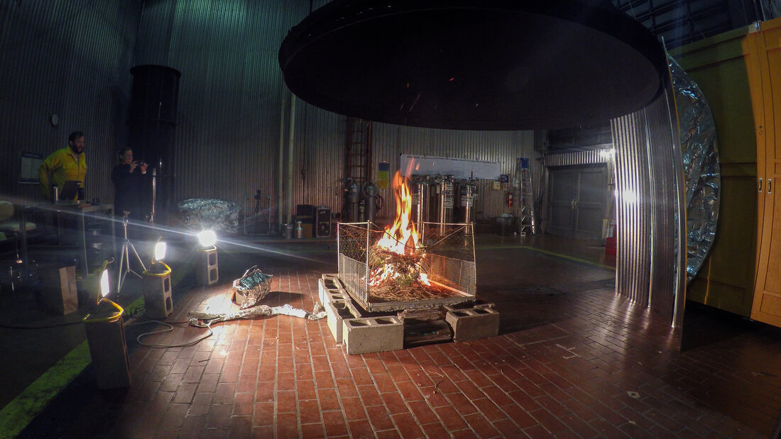 In the middle of a dark room a pile of burning vegetation sends sparks up into a huge, circular vent hood 