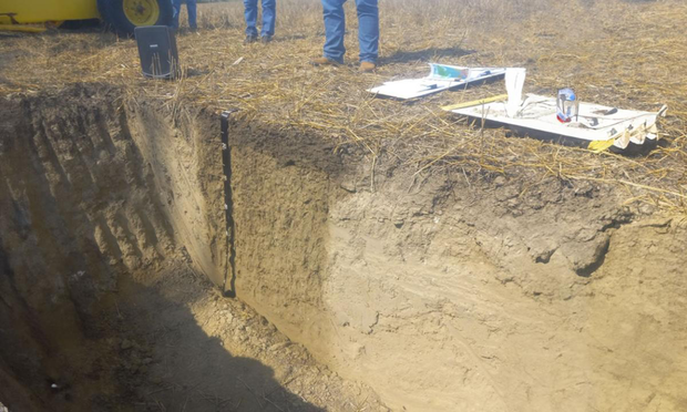 A pit with a vertical wall dug into soil to expose the layers. Instruments and leg + feet of people are on the side.